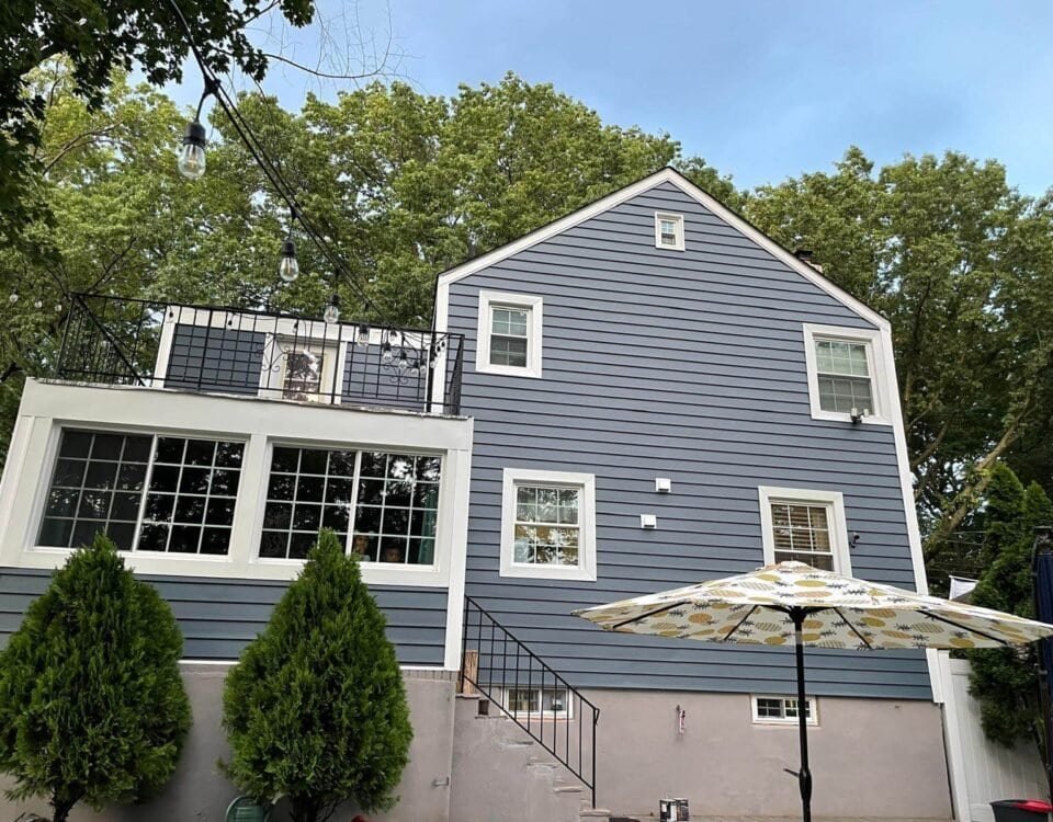 A two-story house with blue siding and white trim, featuring large windows, a black railing on the upper balcony, a staircase, and two green shrubs in front. Perfect for NJ living or anyone seeking Home Remodeling in Union County. A patterned patio umbrella sits below.