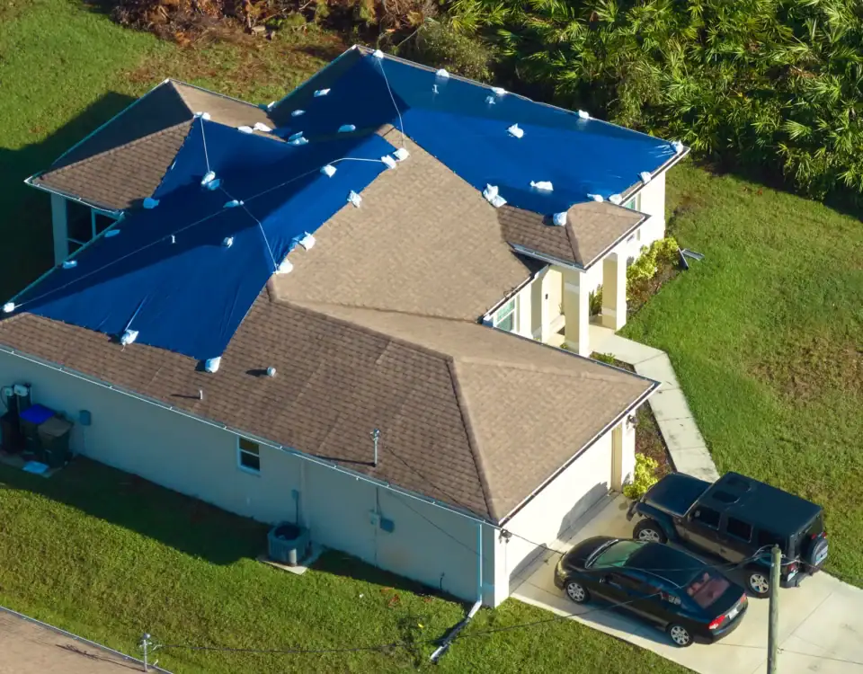 Aerial view of a house with blue tarps covering roof sections, possibly for storm damage repairs or home remodeling in Union County, NJ. Two cars are parked in the driveway, surrounded by green grass and trees.