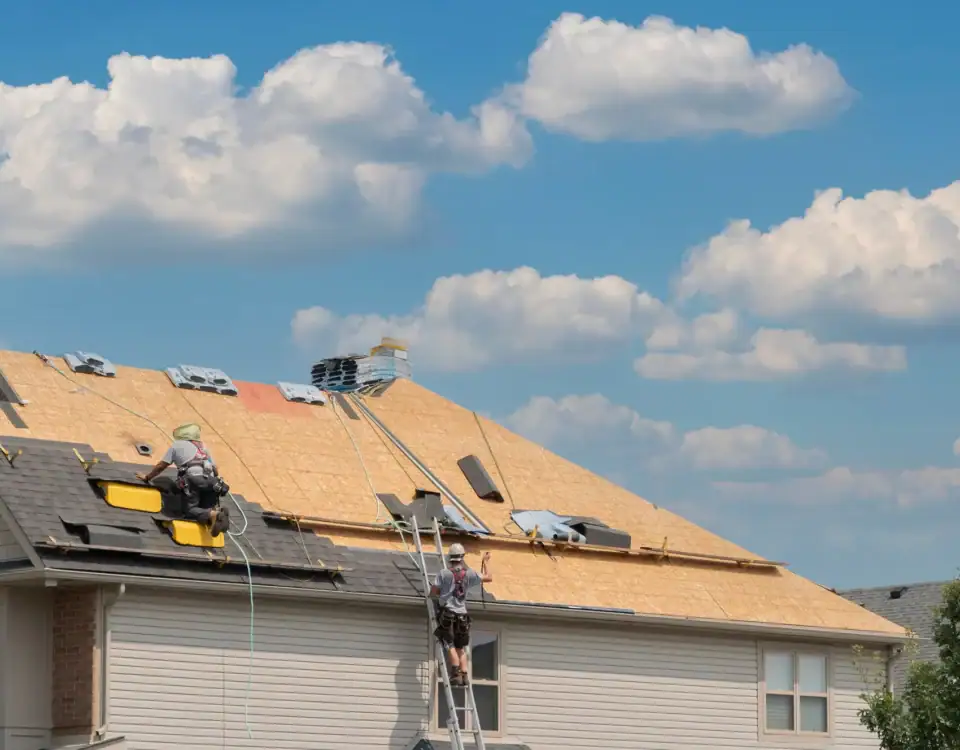 Two workers are installing roofing materials on a house in NJ under a blue sky with scattered clouds. The roof is partially covered with plywood, and equipment is scattered around as one worker climbs a ladder and another sits on the roof.