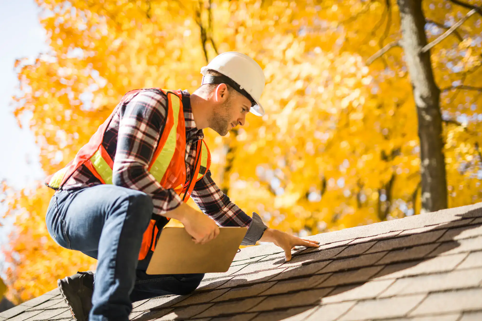 A construction worker in a hard hat and safety vest inspects a roof, clipboard in hand. Vibrant autumn trees fill the background—an example of expert home remodeling in Union County, NJ.