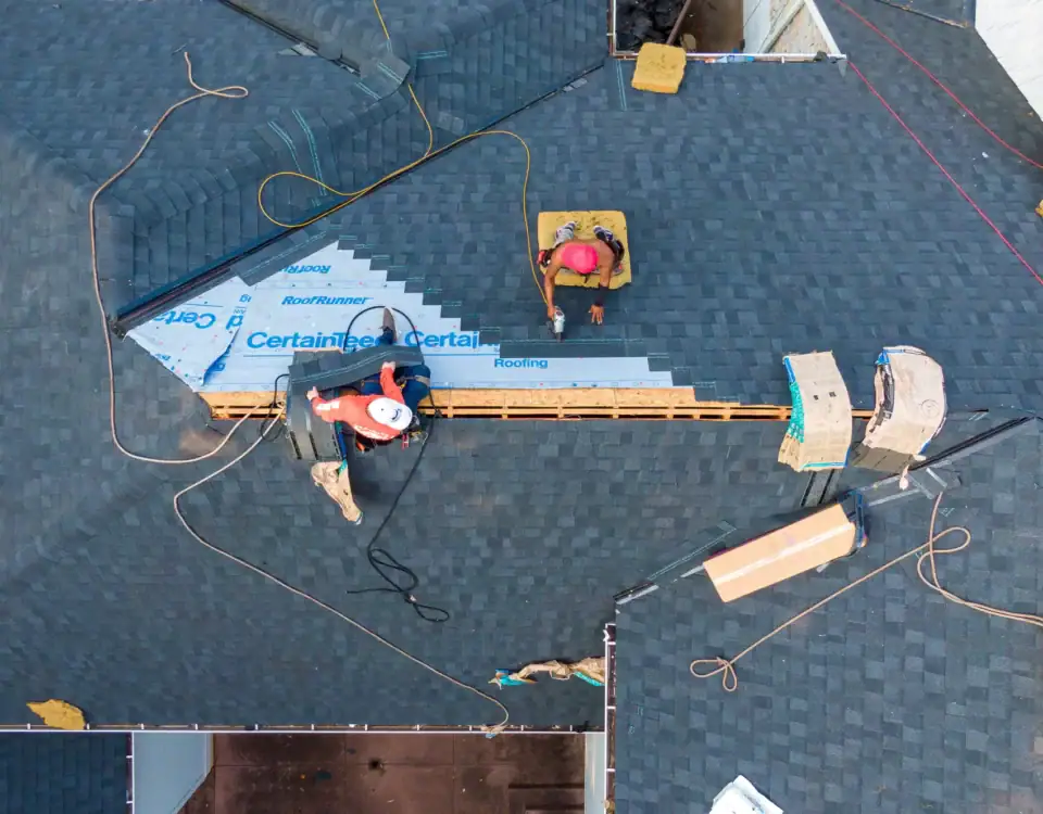 Aerial view of three workers installing shingles and roofing materials on a house roof in Union County, NJ, with tools, hoses, and packaging scattered around during a home remodeling project.