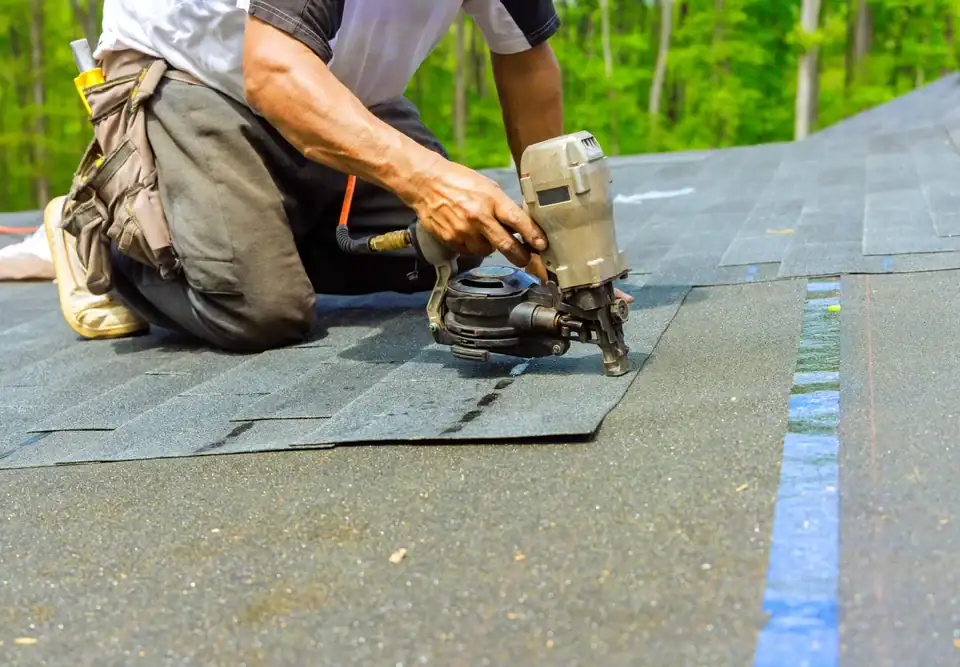 A person kneels on a roof in NJ, using a nail gun to secure asphalt shingles during a roofing installation. Trees are visible in the background, showcasing expert home remodeling in Union County.