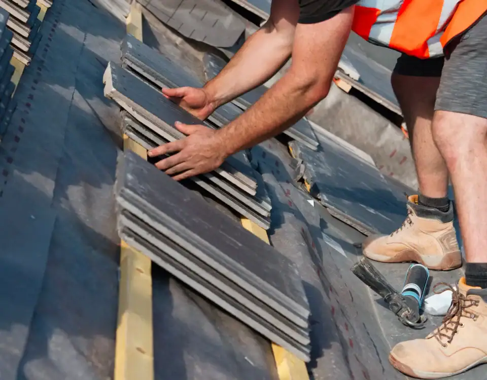 A construction worker wearing boots, an orange safety vest, and shorts installs rectangular tiles on a slanted roof. Hands are placing tiles in neat rows; tools are visible nearby—a common scene in home remodeling in Union County, NJ.