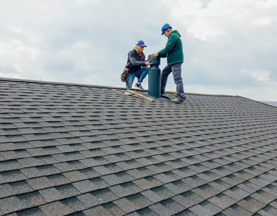 Two workers in blue caps repair or install a vent on a gray shingled roof under cloudy skies, with tools scattered nearby. The scene suggests roofing or maintenance work, possibly part of home remodeling in Union County, NJ.