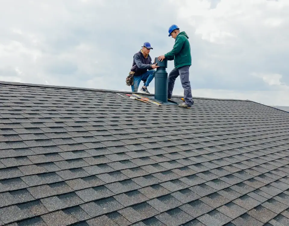 Two workers wearing blue caps are installing or repairing a vent pipe on a shingled house roof, with tools scattered nearby under a cloudy sky—typical of expert home remodeling in Union County, NJ.