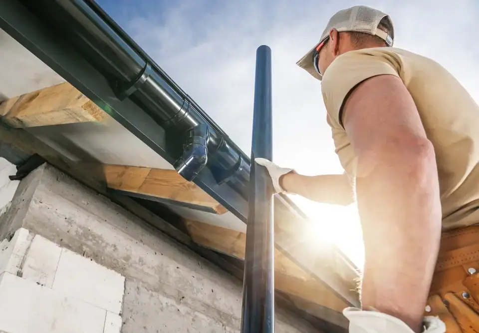 A construction worker wearing a cap, gloves, and tool belt installs or repairs a rain gutter system on the edge of a building roof under bright sunlight, showcasing expert Home Remodeling Union County skills in NJ.