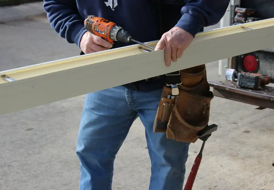 A person in jeans and a tool belt uses a power drill to screw into a long piece of wood, working outdoors on a concrete surface.