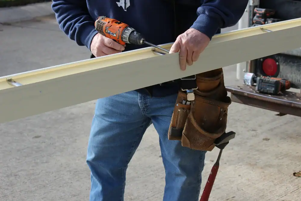 A person in jeans and a tool belt uses a power drill to screw into a long piece of wood, working outdoors on a concrete surface.