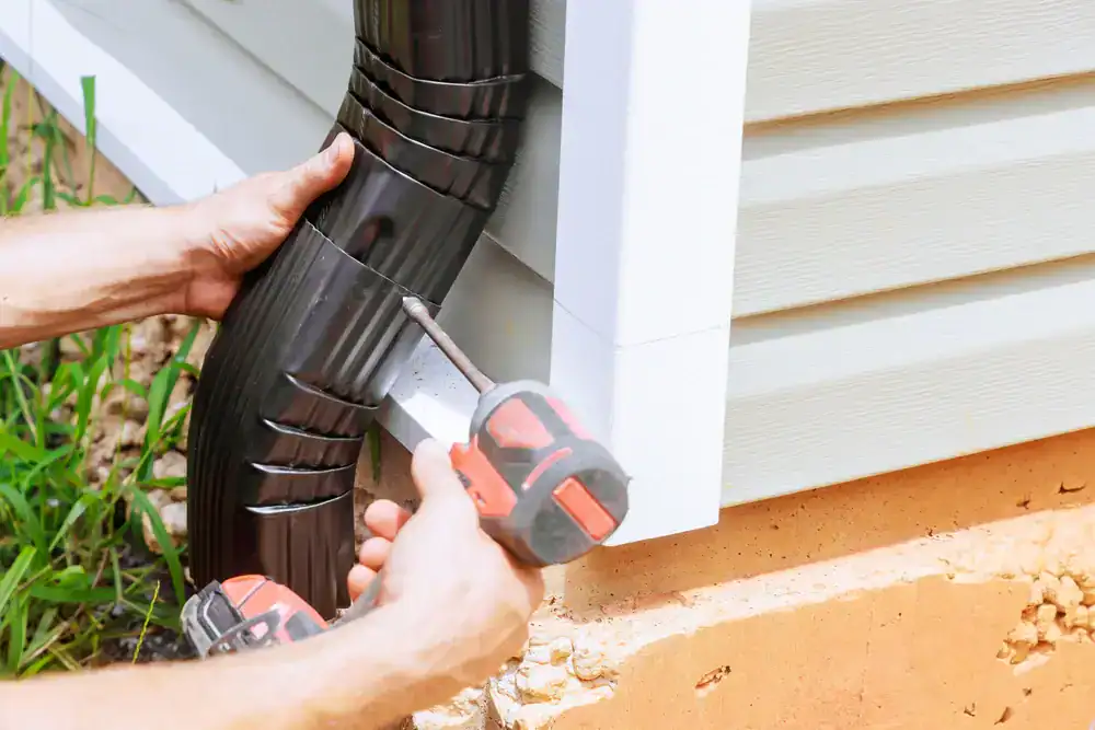 A person uses a cordless power drill to attach a black downspout to the side of a house with beige siding and a concrete foundation, showing the quality workmanship offered by Roofing Services Union County, NJ.