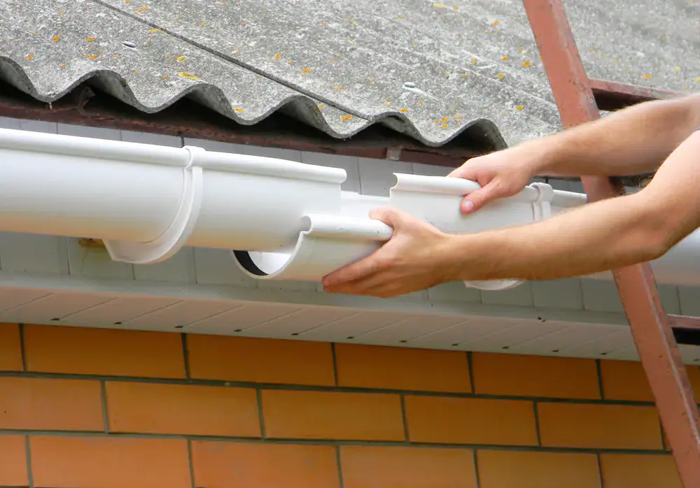A person installs or repairs a white plastic rain gutter on the edge of a roof with corrugated sheets above a brick wall, using both hands while standing on a ladder.