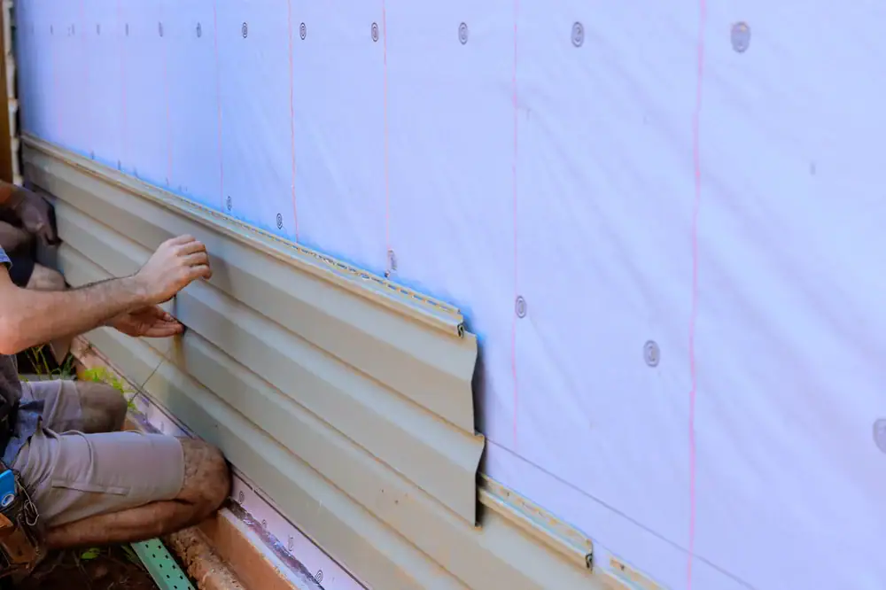 A person installs beige vinyl siding panels on the exterior wall of a house covered with a white weather-resistant barrier.