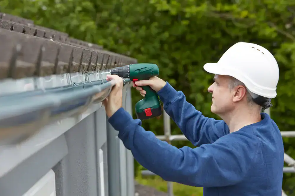 A person wearing a white hard hat and blue sweatshirt uses a green cordless drill to install or fix a rain gutter on the edge of a building roof in NJ, with trees visible in the background. Roofing Services Union County can help with similar projects.
