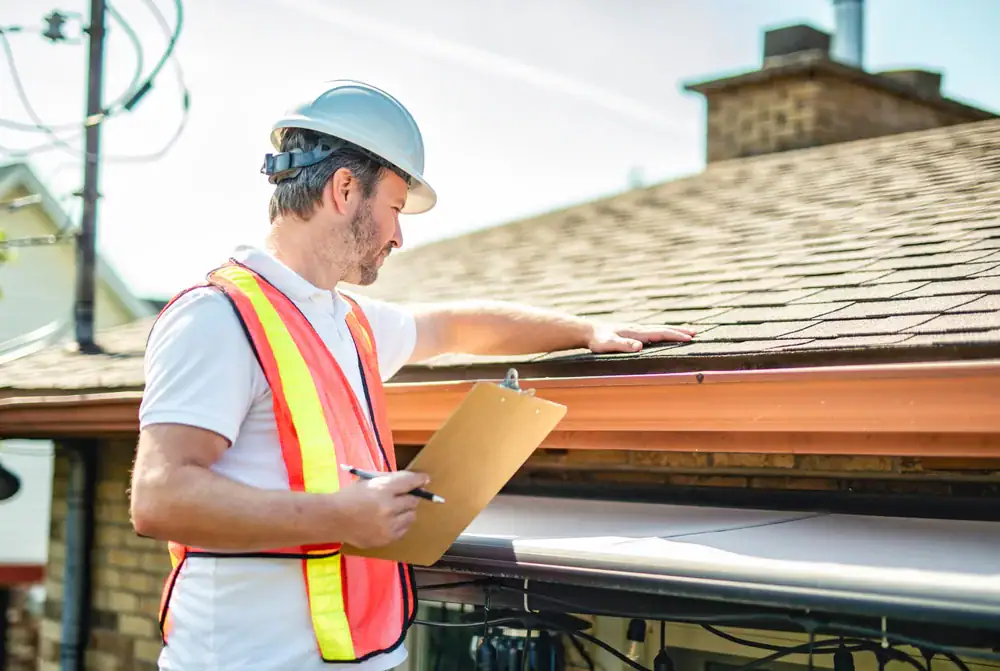 A man wearing a hard hat and reflective safety vest inspects the shingles on a residential roof while holding a clipboard and pen.
