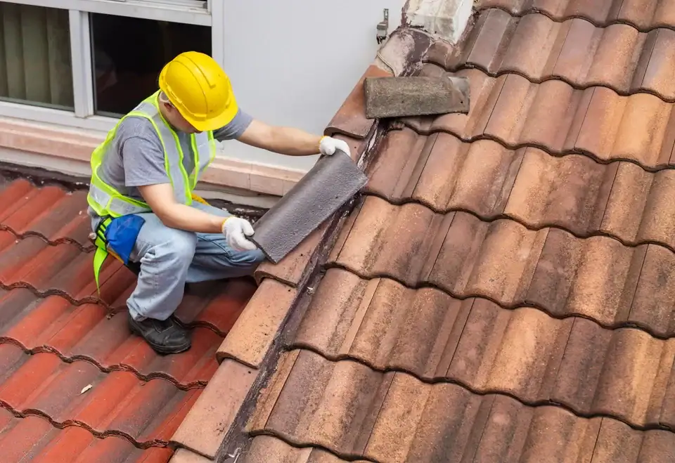 A construction worker wearing a yellow hard hat and safety vest repairs a tiled roof, placing new material along the roof valley near a window.