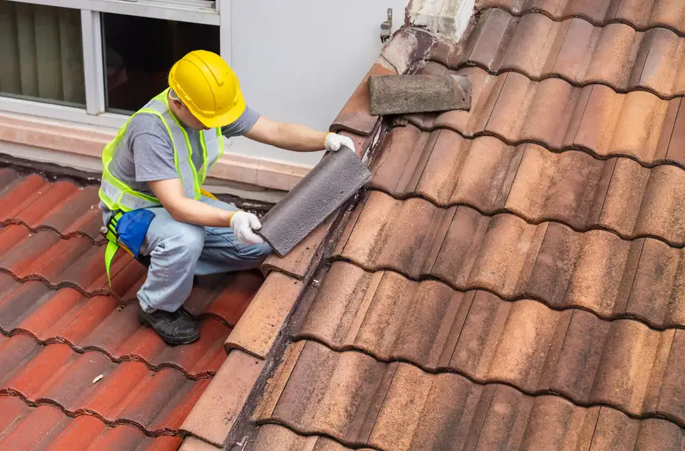 A construction worker wearing a yellow hard hat and safety vest repairs a tiled roof, placing new material along the roof valley near a window.