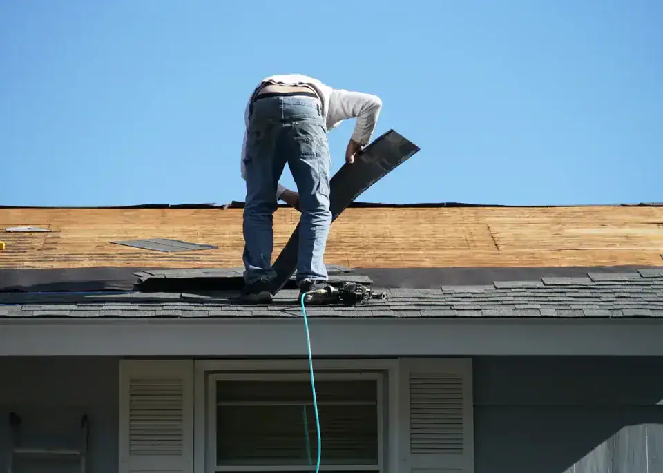 A person stands on a house roof in NJ installing or repairing shingles under a clear blue sky, with work tools and materials visible nearby—a typical scene for Roofing Services Union County professionals.