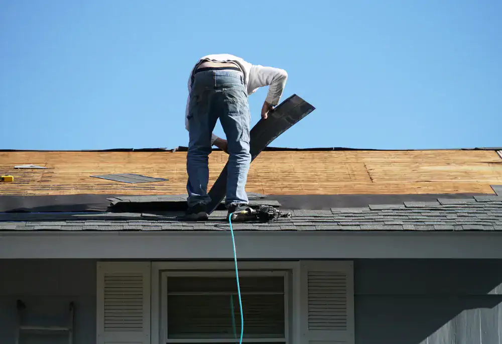 A person stands on a house roof in NJ installing or repairing shingles under a clear blue sky, with work tools and materials visible nearby—a typical scene for Roofing Services Union County professionals.