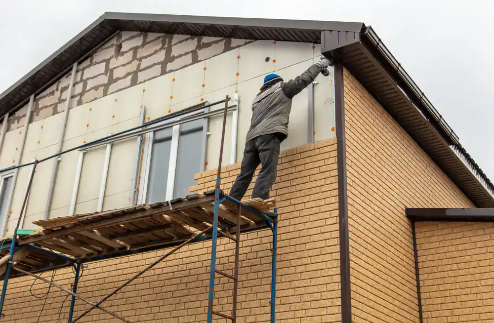 A person standing on scaffolding installs siding on the upper exterior of a two-story brick house under construction or renovation. The worker, equipped with protective clothing and a helmet, exemplifies the quality of Roofing Services Union County, NJ.