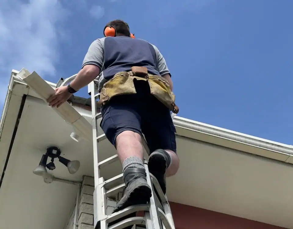 A person wearing a tool belt, work boots, and ear protection climbs a ladder to perform maintenance on the roofline of a house under a clear blue sky.