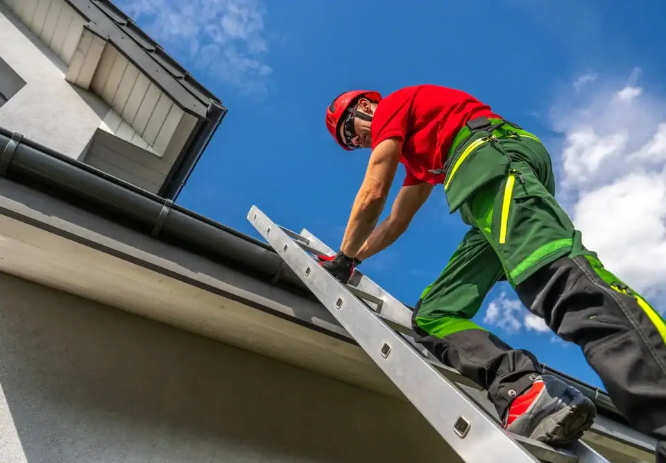A worker in safety gear climbs a ladder to inspect or repair a house roof and gutter on a sunny day, with the blue sky in the background.