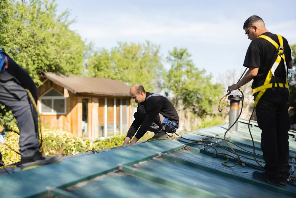 Three workers in safety gear install or repair a metal roof on a sunny day, with trees and a wooden shed in the background. One worker crouches to adjust the roof while another holds equipment.