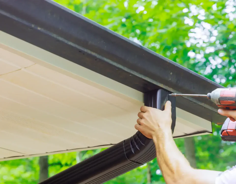 A person uses a power drill to attach a black gutter to the edge of a roof, surrounded by green trees in the background, showcasing expert Roofing Services in Union County, NJ.