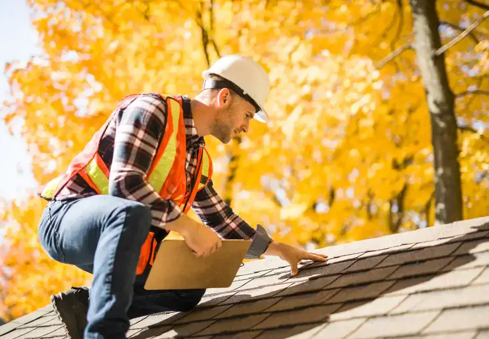 A construction worker wearing a white hard hat, orange safety vest, and plaid shirt inspects a shingled roof with a clipboard in hand. Bright yellow autumn trees are visible in the background, showcasing expert Roofing Services Union County,NJ.
