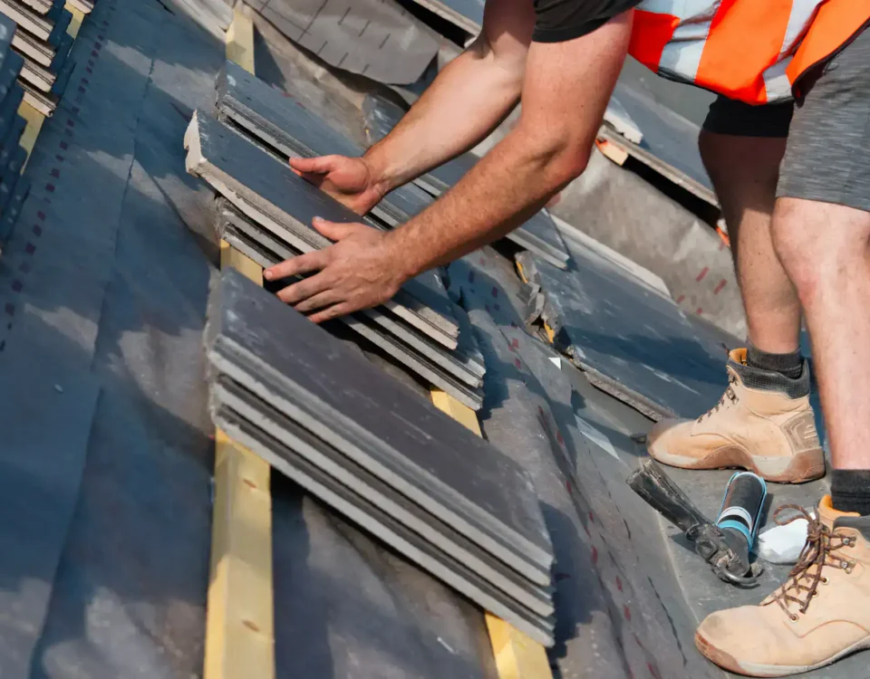 A worker in boots and an orange safety vest is installing gray roof tiles on a sloped roof, using both hands to position the tiles carefully—a scene typical of Roofing Services Union County, NJ.