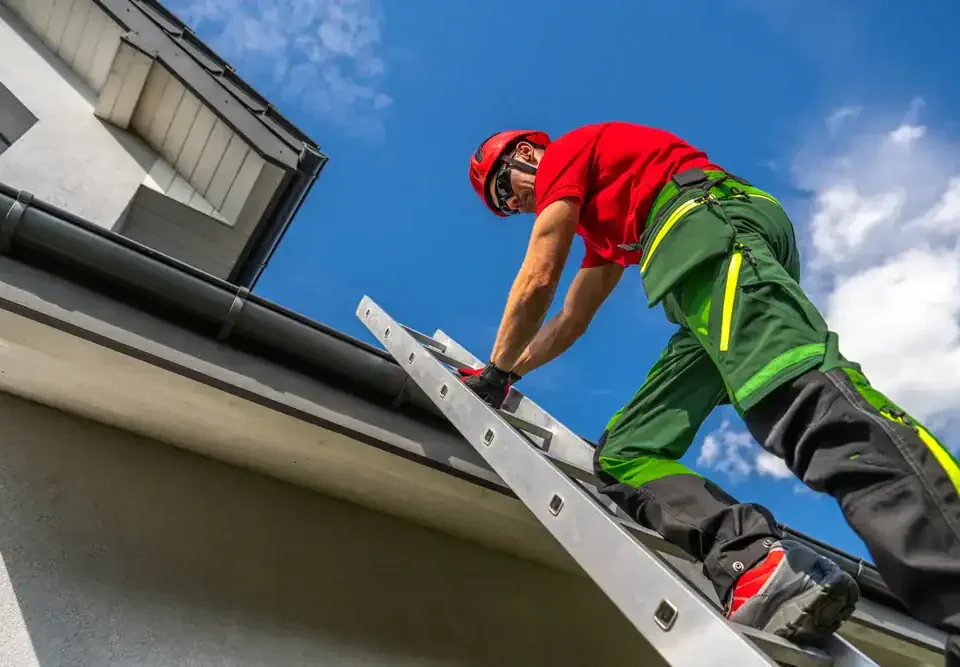 A worker in safety gear climbs a ladder to reach the roof of a house under the blue NJ sky. Wearing a red helmet, red shirt, and green pants, he ensures proper safety while providing Roofing Services Union County residents trust.