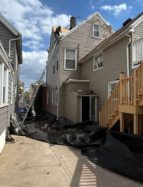 A tan house with damaged roof shingles and debris covering the ground beside it, likely from a storm. The sky is partly cloudy, and the neighboring house is visible on the left side.