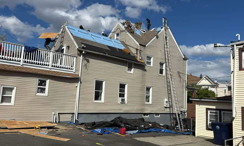 Workers repair the roof of a beige house using ladders and blue tarps, with materials and tools scattered on the ground and deck. A partly cloudy sky appears overhead.