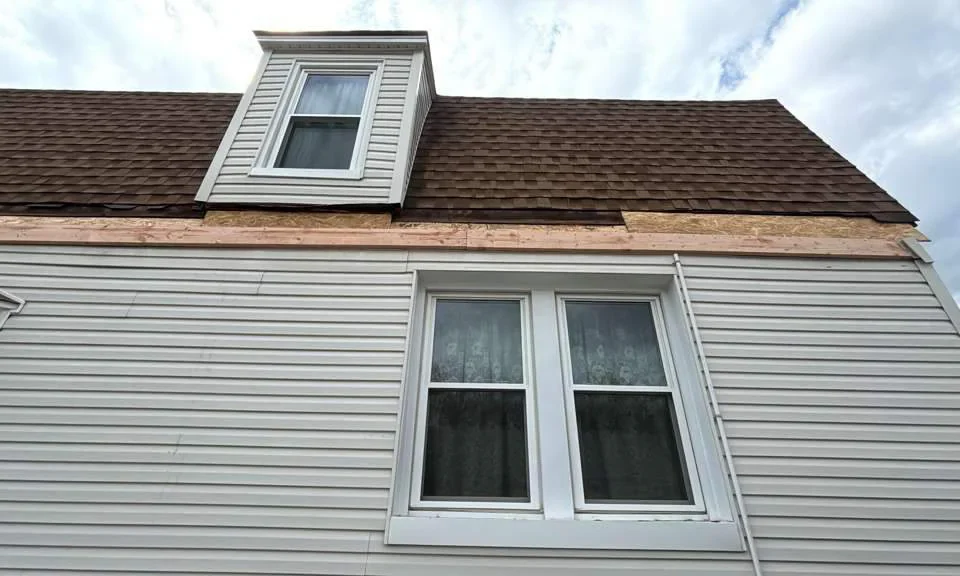 A house exterior showing white siding, a double window on the lower floor, and a dormer window above. There’s unfinished wood trim beneath the brown shingled roof, indicating ongoing construction or repairs.