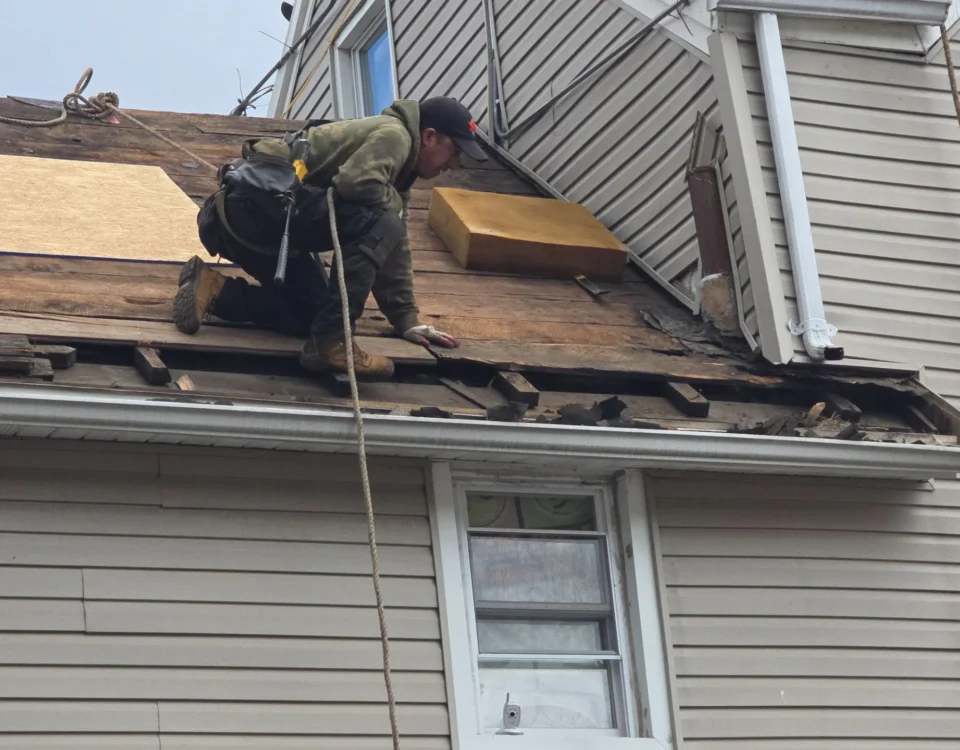 A construction worker wearing safety gear kneels on a sloped wooden roof, repairing damaged boards on a house. Tools and materials are scattered nearby. The roof's shingles have been removed.