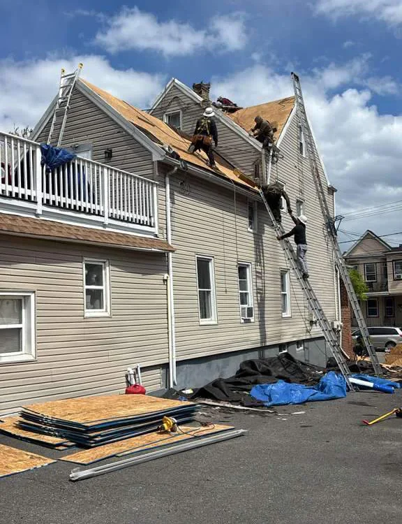 Three workers repair or install roofing on a two-story house with beige siding. Ladders, roofing materials, and tools are visible on the ground, and the sky is partly cloudy above.