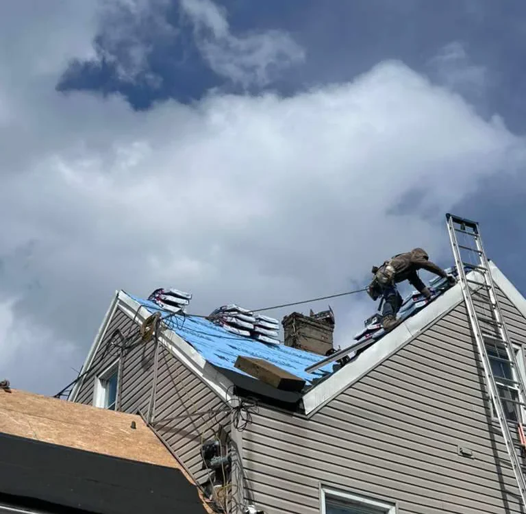 A worker repairs the roof of a house, kneeling on blue tarp and tethered to a safety line. Roofing materials are stacked nearby, and a ladder is propped against the steep roof under a partly cloudy sky.