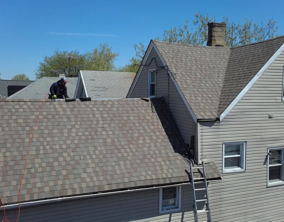 A person works on the roof of a beige house using tools, with a ladder propped against the side. The sky is clear and blue, and nearby trees have green leaves.