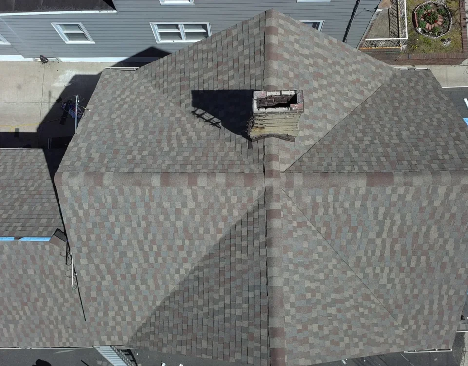 Aerial view of a shingled rooftop with a central chimney. Part of the roof is under repair, with workers and roofing materials visible on the left side. A nearby house and yard are partially visible.
