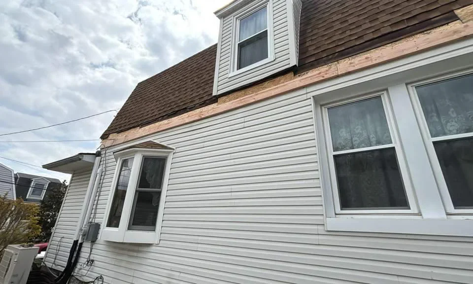 A white house with brown shingles is shown. The siding near the roof is removed, exposing wood underneath. There are several windows, including a bay window and an upper dormer window. The sky is partly cloudy.